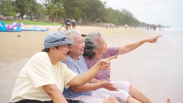 Happy senior women friends travel at the sea, sitting and celebrate aging gracefully together at the beach, active elderly retirement people lifestyle, female friendship on summer holiday vacation.