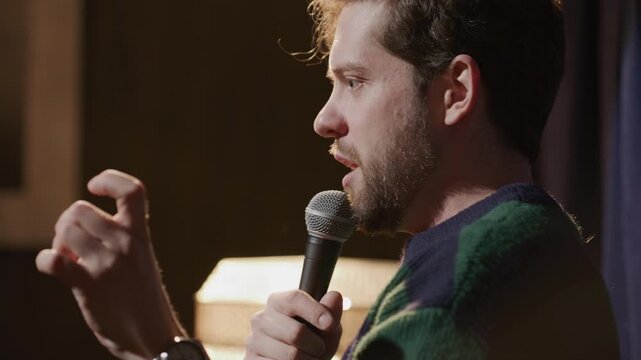 Side view of young man speaking into microphone, telling story on warmly lit stage during live stand up performance at comedy club