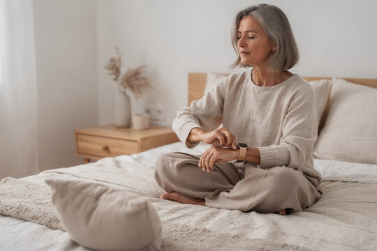 Peaceful senior woman sitting cross-legged on a bed in a minimalist bedroom, checking her fitness stats on a smartwatch