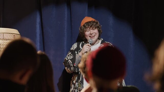 Young female standup comedian in stylish outfit sharing hilarious story and laughing on stage during live performance at comedy club