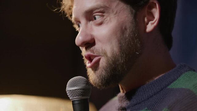 Cheerful male comic smiling while telling witty story into microphone during standup show on stage. Close up view