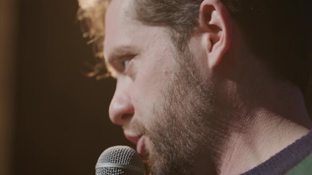 Young male stand up performer delivering speech with microphone on stage illuminated by warm light during comedy show. Close up shot
