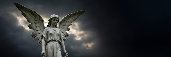 Fototapeta premium Weathered stone angel statue with large wings stands against dramatic dark sky. Spiritual concept of grief, faith, and mourning. Panoramic banner with copy space