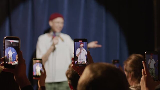 Audience holding smartphones while recording female comedian performing stand up show on stage, camera focusing on device screens capturing live comedy set