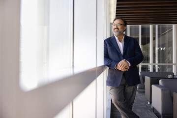 Fototapeta premium Smiling businessman leader stands by office window, reflecting on goals, ideas or upcoming decisions during break, represents strategic thinking, planning and professional confidence. Business vision