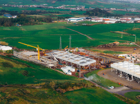Aerial view of the Pico Vermelho Geothermal Power Station under construction with a yellow crane and industrial cooling units surrounded by green hills and fields in Conceicao, Acores, Portugal.