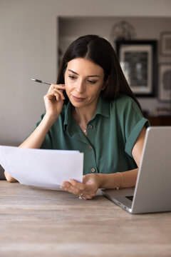 Focused adult woman sits at desk, reading documents text, analyzing details, reviewing information of contract terms data, learning insurance papers, doing remote work from home office, vertical shot
