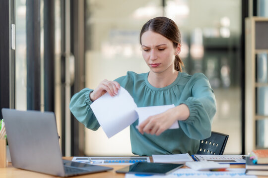 Angry businesswoman tearing up paper document at office desk with frustrated expression.