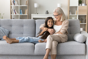 Obraz premium Loving older grandmother cuddling cute little granddaughter, smile, lead trustworthy conversation, give advice, express care, share support on weekend leisure at home. Multigenerational family ties