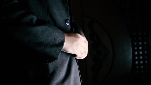 Close up of a man in a dark tailored suit buttoning his jacket in dramatic cinematic low light. Set against a pitch-black background, the low-key lighting creates a moody and mysterious atmosphere.