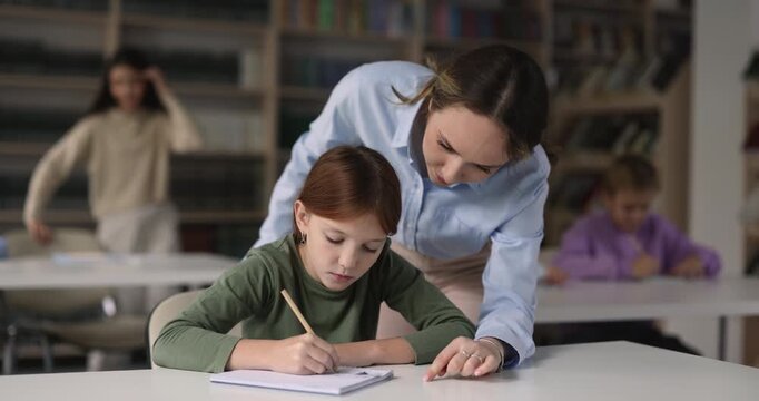Female educator guiding to schoolgirl learning, making schoolwork in copybook sit at desk during lesson in classroom. Primary school education, schooling, knowledge and skills development of child