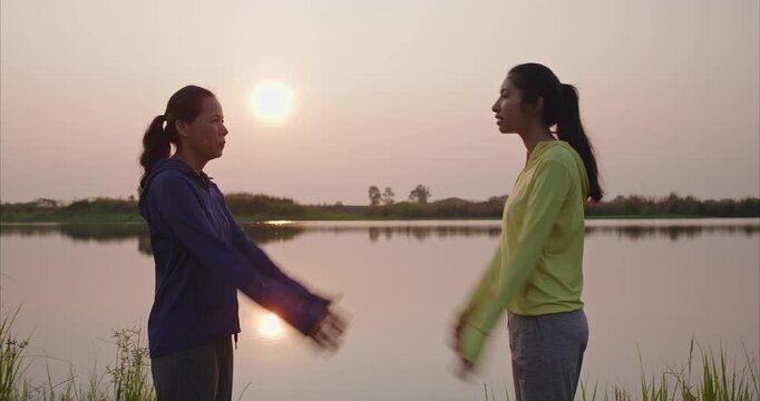 healthy Asian mother and daughter practicing traditional swinging arm exercise together for holistic wellness at the tranquil lakeshore during a beautiful golden hour sunset.