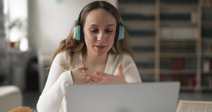 Female university student in headphones speaking with online tutor or classmate, discussing assignment using video call application on laptop in library. Remote education, digital communication, study