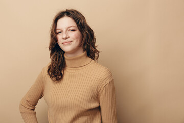 Obraz premium Young woman with curly brown hair wearing beige turtleneck sweater smiles confidently against plain beige background. Concept of casual style, natural beauty, and positive mood.