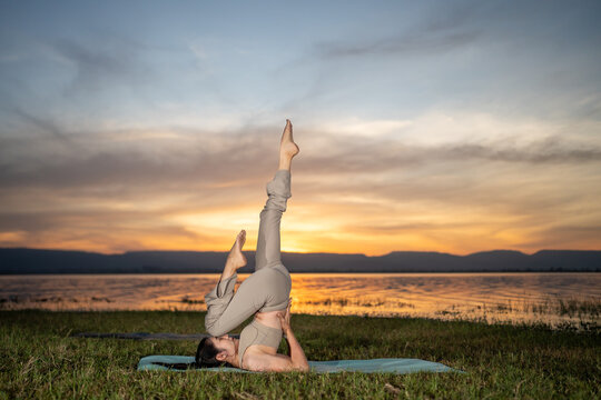 Young woman practicing shoulder stand yoga pose outdoors by a peaceful lake at sunset. Concept of flexibility, balance, mindfulness, wellness, and healthy lifestyle in natural scenery.