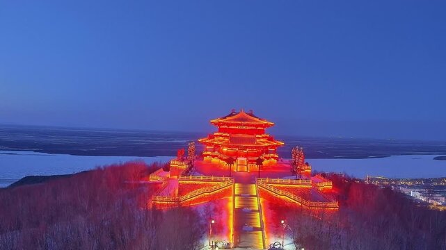 A spectacular brightly lit red traditional pavilion on a hill overlooking the frozen river at night in Heilongjiang.