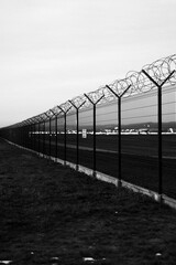 Fototapeta premium Dramatic black and white vertical shot of a high-security metal fence topped with razor wire coils, bordering a restricted airport runway area under a moody, overcast evening sky.