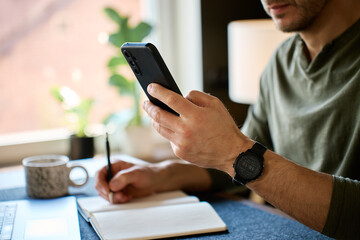 Fototapeta premium Man checking smartphone and writing in notebook at desk with laptop in home office. Concept of daily planning, digital assistant, remote work and everyday technology