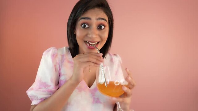 Close up of happy smiling beautiful young Indian woman lady hold glass enjoying delicious orange juice looking at camera feels surprised, isolated against pink studio background.