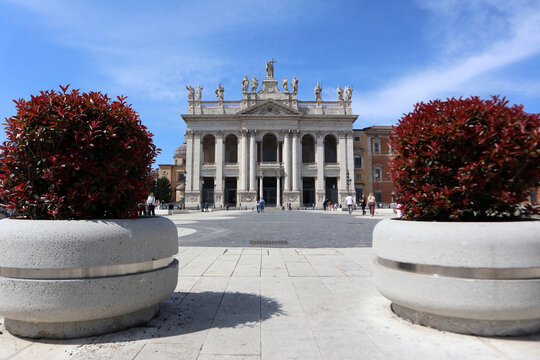 Facciata monumentale dell'Arcibasilica di San Giovanni in Laterano a Roma, vista prospettica dalla piazza con grandi fioriere moderne in primo piano e cielo blu