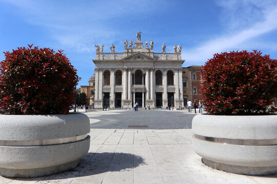 Facciata monumentale dell'Arcibasilica di San Giovanni in Laterano a Roma, vista prospettica dalla piazza con grandi fioriere moderne in primo piano e cielo blu