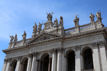 Obraz premium Facciata monumentale dell'Arcibasilica di San Giovanni in Laterano a Roma, vista prospettica dalla piazza con grandi fioriere moderne in primo piano e cielo blu