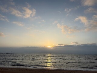 Obraz premium Cinematic landscape showing the sun rising above a low layer of clouds over a calm sea, with sand and a distant lighthouse silhouette.