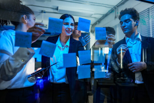 Diverse professional team engaged in a high energy brainstorming session, using sticky notes on a glass wall in a modern office at night.