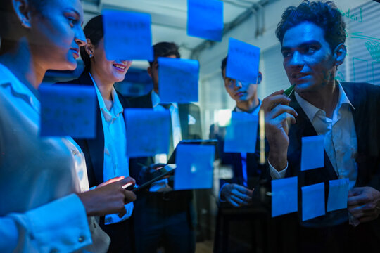 Diverse cyber security team mapping out a secure network defense strategy on a glass wall in a high tech corporate office at night.