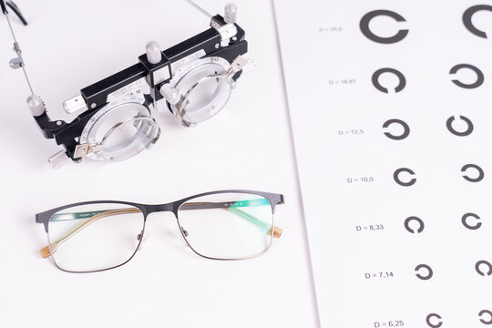 White background features a clear eye chart with decreasing letter sizes. Two eyeglasses sit beside an eye measurement device. One has a dark frame, the other metal