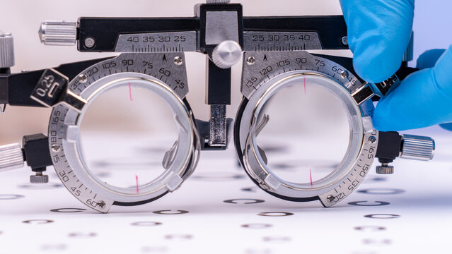 Close-up of a phoropter for eye examinations. Gloved hand adjusts the device's lenses. The background shows an eye chart with symbols. This equipment helps measure vision accuracy