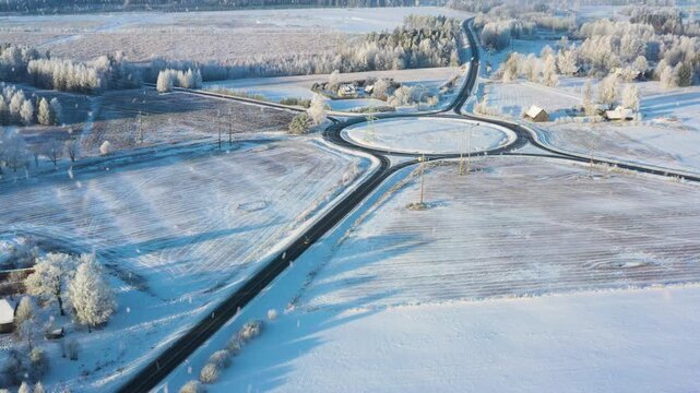 Roads converging at a winter roundabout, vehicles navigating the snow covered landscape with snowfall in aerial view