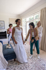 Naklejka premium Female bridal fitting at home, bride in gown, Asian fitter measuring hem with tape, senior watching