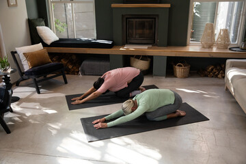 Naklejka premium Female friends practicing yoga on two black mats in living room by fireplace and bench