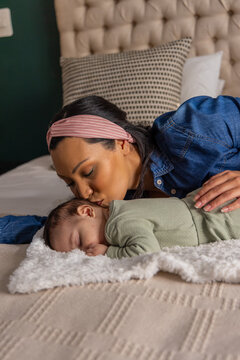Kissing mother leaning over sleeping infant on bed, wearing denim shirt and pink striped headband