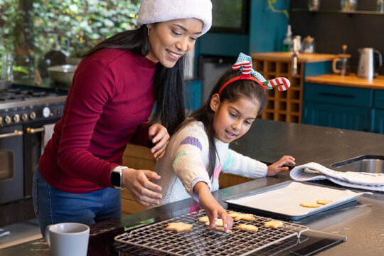 Asian mother in white hat guiding daughter placing star cookies on cooling rack at kitchen island