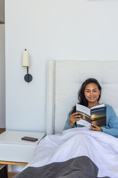 Adult Asian woman sitting in bed reading Endless Sunsets book with phone on nightstand, copy space