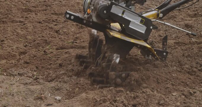 Close-up of a rotary cultivator tilling the soil, preparing the land for planting seeds in a garden. The machine's blades spin, digging into the earth and turning it over for farming work.