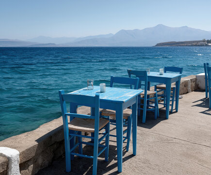  Iconic blue wooden chairs and tables sit empty beside turquoise calm Aegean waters.