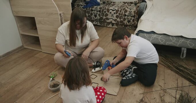 Creative family time. Mother, son, and daughter are sitting on the floor, happily crafting with natural materials like twigs and using glue guns for their DIY projects. The family is making a craft.