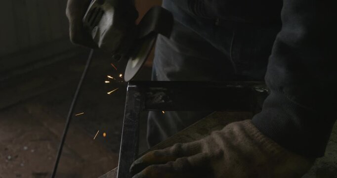 Professional metalworker in protective gloves using an angle grinder to clean a metal profile pipe, creating a shower of bright sparks in a dimly lit workshop before welding.