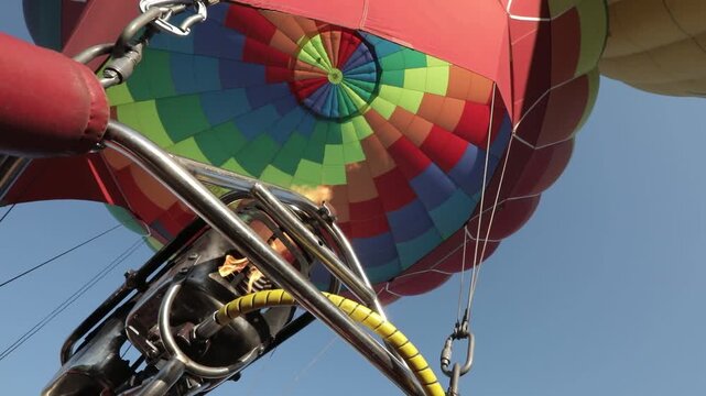 A vibrant hot air balloon with a colorful pattern is being inflated under a clear blue sky, viewed from the basket.