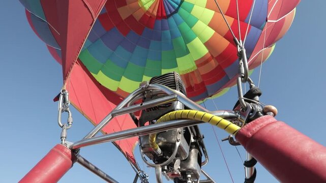 A vibrant hot air balloon with a colorful pattern is being inflated under a clear blue sky, viewed from the basket.