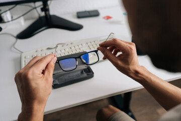 Over the shoulder view of man holding eyeglasses with blue light filter. Person taking break from...