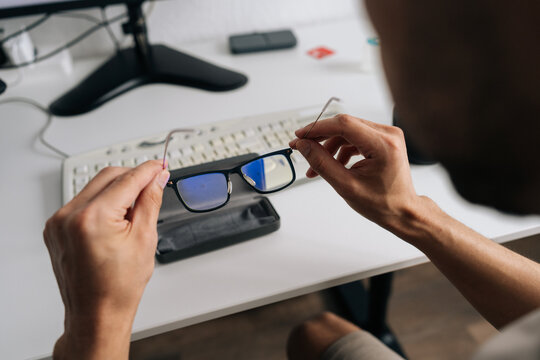 Over the shoulder view of man holding eyeglasses with blue light filter. Person taking break from computer work to prevent eye strain. Modern office home office setting with white desk keyboard.