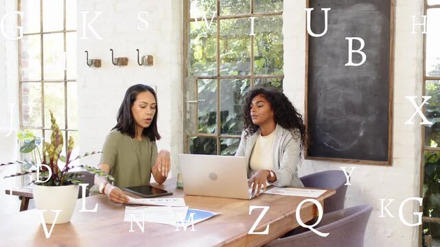Left woman starting design review, handing chart amid text, right peer typing, showing laptop