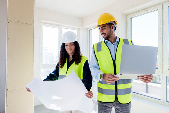 Architect holding laptop with colleague reviewing blueprint at site