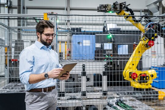 Smiling engineer standing and using tablet PC near robotic arm at factory