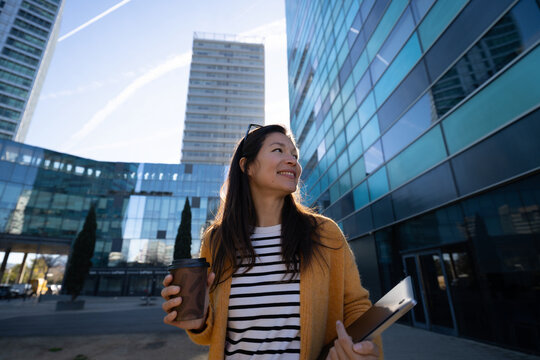 Smiling businesswoman holding coffee and laptop in modern city