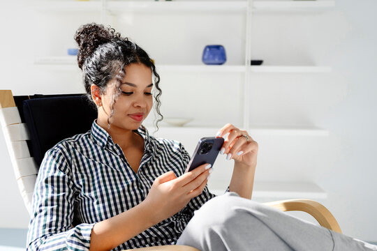 Curly haired model relaxing indoors texting on smartphone